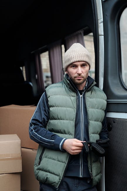 A man wearing a light grey knit beanie, a dark navy jacket with blue accents, and a green padded vest standing inside the back of a moving van. He is holding a black strap used for securing furniture or boxes during a home relocation. Behind him, there are stacked cardboard moving boxes of various sizes. The interior of the van appears well-lit, with a glimpse of the vehicle's window and curtain visible in the background. The scene captures the process of packing and loading items for a furniture transport or house removal service, illustrating prepared packing materials and careful handling, as provided by West Ealing Removals.