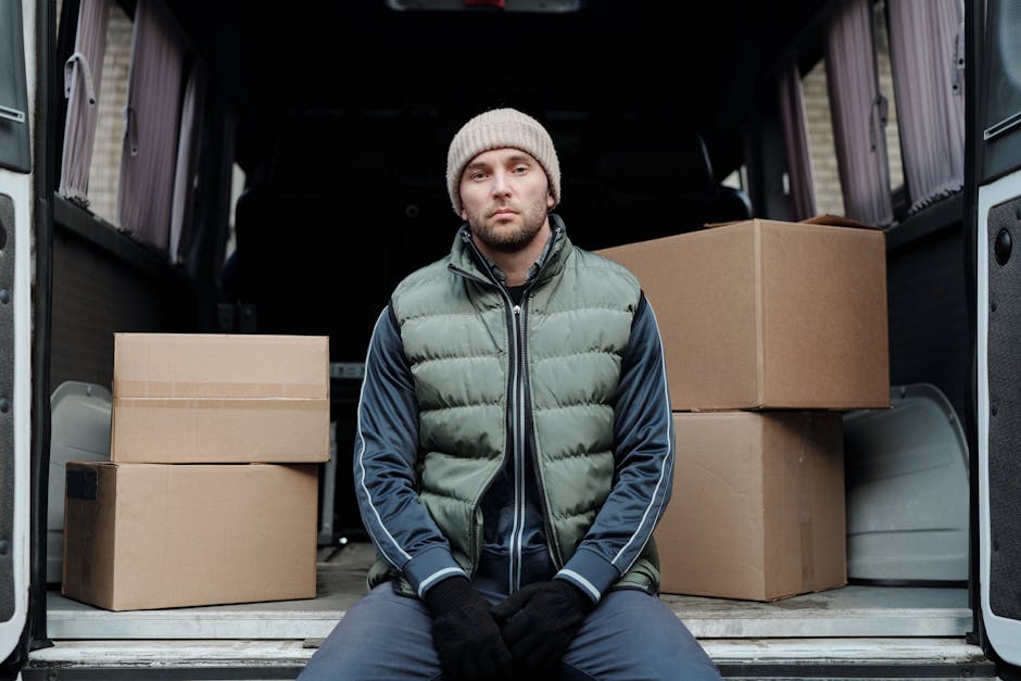 A young man with light skin, a neutral expression, and a beard is seated inside the open cargo area of a moving van, which contains several cardboard boxes of various sizes, some sealed with packing tape. The boxes are stacked on the vehicle's floor, which is made of ribbed metal, and a few are resting against the van's interior walls. The man is dressed in a beige knit beanie, a green puffer vest layered over a navy zip-up jacket with white stripes along the sleeves, and black gloves. The environment appears to be outdoors, with natural lighting illuminating the scene, and the background suggests a residential area. This image depicts the process of home relocation, with the individual possibly involved in moving or packing furniture and possessions, supported by professional removal services such as those offered by West Ealing Removals.