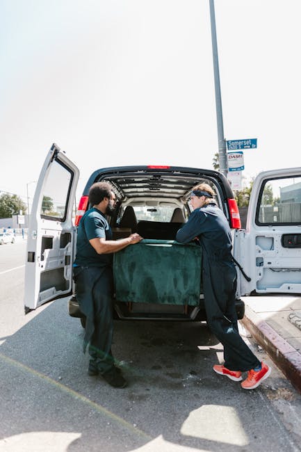 A man wearing a light grey knit beanie, a dark navy jacket with blue accents, and a green padded vest standing inside the back of a moving van. He is holding a black strap used for securing furniture or boxes during a home relocation. Behind him, there are stacked cardboard moving boxes of various sizes. The interior of the van appears well-lit, with a glimpse of the vehicle's window and curtain visible in the background. The scene captures the process of packing and loading items for a furniture transport or house removal service, illustrating prepared packing materials and careful handling, as provided by West Ealing Removals.