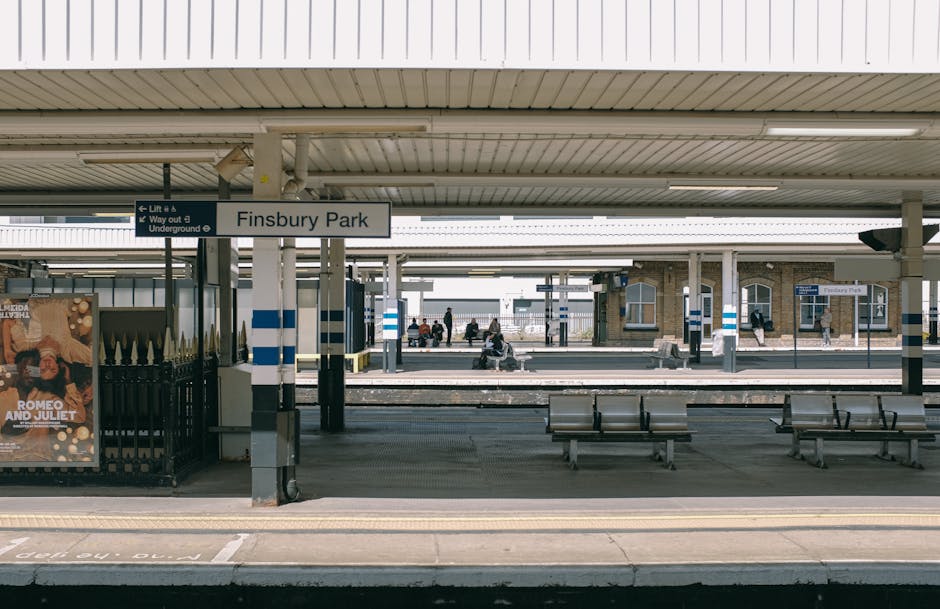 The image depicts a view of Finsbury Park railway station platform with a white and blue sign displaying the station name. The platform is covered by a metal roof with fluorescent lighting, supported by columns. Several people are seated on benches along the platform, some waiting while others are walking or standing near the edge. A poster advertising 'Romeo and Juliet' is mounted on a black iron fence to the left. In the background, tracks are visible along with additional platforms, station buildings, and some passengers. The overall scene is well-lit with natural daylight, portraying a typical urban train station environment suitable for house removals and furniture transport logistics in the Ealing area, connected to West Ealing Removals' services.