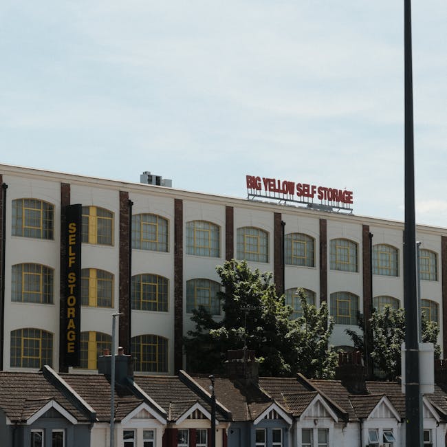 Exterior view of a multi-story building with a sign reading 'BIG YELLOW SELF STORAGE' in red letters on the roof and a vertical yellow sign on the side stating 'SELF STORAGE.' The building has large, arched windows with yellow frames, and there are several trees partially obscuring the lower part of the structure. In the foreground, there is a row of small houses with sloped roofs and chimneys, situated along the street. The scene is taken during daytime with clear weather, and part of a streetlamp is visible on the right side of the image. This environment depicts a commercial self-storage facility likely used for home relocation and furniture transport services, aligned with house removals and moving logistics.