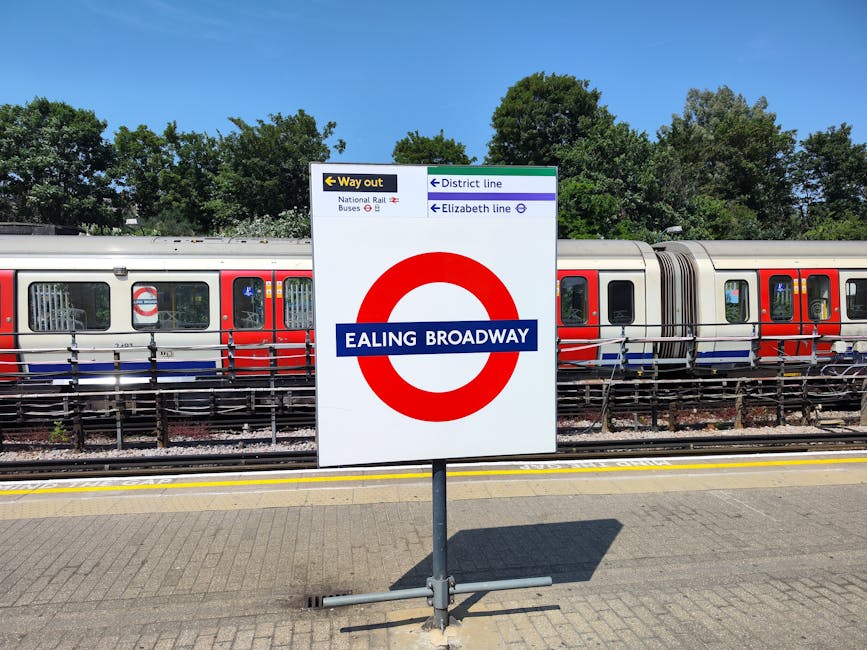 The image depicts a view of Finsbury Park railway station platform with a white and blue sign displaying the station name. The platform is covered by a metal roof with fluorescent lighting, supported by columns. Several people are seated on benches along the platform, some waiting while others are walking or standing near the edge. A poster advertising 'Romeo and Juliet' is mounted on a black iron fence to the left. In the background, tracks are visible along with additional platforms, station buildings, and some passengers. The overall scene is well-lit with natural daylight, portraying a typical urban train station environment suitable for house removals and furniture transport logistics in the Ealing area, connected to West Ealing Removals' services.